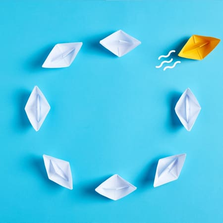A group of seven white paper boats arranged in a circular formation on a blue background. An eighth paper boat, colored yellow, is outside the circle with three wavy lines near it, indicating movement away from the group.