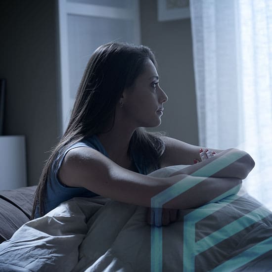 A woman with long brown hair sits in bed with light streaming in through a window. She looks contemplative, gazing outwards. She is wrapped in a white comforter, and the surroundings suggest a calm, early morning atmosphere.