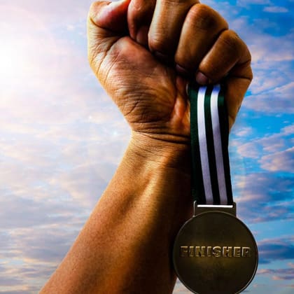 A close-up of a raised fist holding a "FINISHER" medal with a striped ribbon against a backdrop of a cloudy sky. The sunlight creates a dramatic effect, highlighting the sense of achievement.