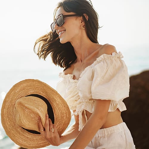 A woman wearing sunglasses and a white off-shoulder blouse smiles while holding a wide-brimmed straw hat. She is near the sea, with the sun casting a warm glow on her. Her hair is flowing in the breeze, and she appears relaxed and happy.