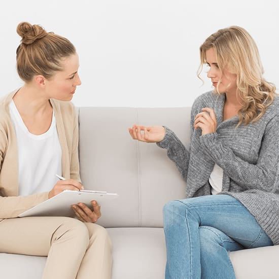 Two women are sitting on a couch facing each other. One woman, holding a clipboard and pen, appears to be a therapist or counselor, while the other woman, who is gesturing with her hands, seems to be speaking or explaining something. Both look engaged in conversation.