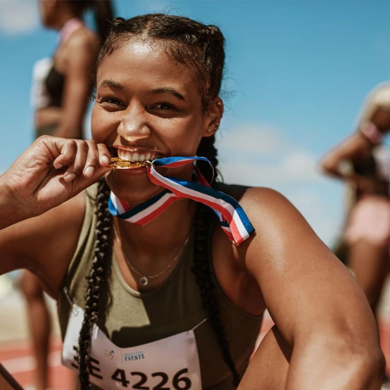 A female athlete with braided hair smiles brightly as she crouches on a track field, biting onto a gold medal. She wears a race bib with the number 4226 and other runners can be seen in the blurred background under a clear blue sky.