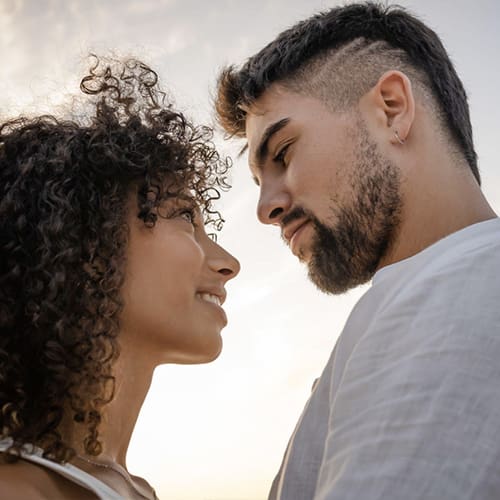 A couple stands closely facing each other against a soft, cloudy sky. The woman on the left has curly hair and smiles, and the man on the right has short, styled hair with an undercut and a beard. They seem to be sharing an intimate moment, looking into each other's eyes.