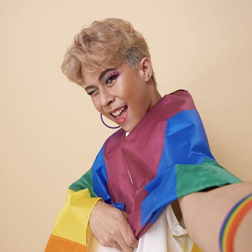 A person with blonde hair and colorful makeup is smiling and winking at the camera. They are draped in a rainbow flag, holding one end of it, and wearing a rainbow wristband. The background is a plain beige wall.