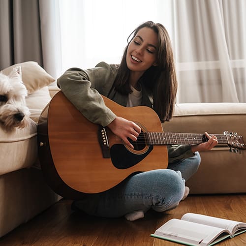 A woman sits on a wooden floor, leaning against a couch, playing an acoustic guitar and smiling. A small dog is resting its head on the couch near her. There's an open book on the floor beside her. Natural light filters through sheer curtains in the background.