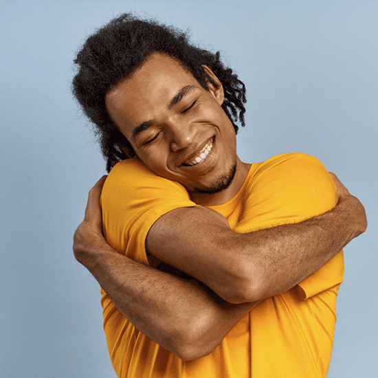 A person with medium-length curly hair wearing an orange shirt is smiling with their eyes closed while hugging themselves. They stand against a plain light blue background.