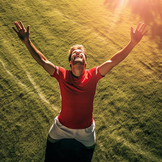 A soccer player in a red shirt and white shorts celebrates on a grass field, arms raised and face turned upwards with a joyful expression. The sunlight illuminates the scene from above, creating a warm and triumphant atmosphere.
