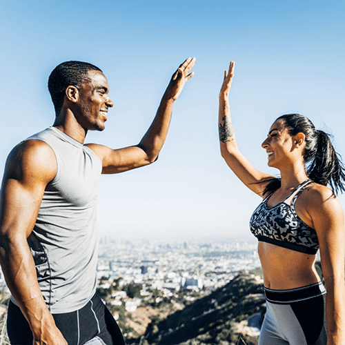 A man and a woman in athletic wear high-five outdoors on a sunny day. The background features a scenic view of a city skyline and rolling hills. They appear happy and energetic, suggesting a successful exercise session or hike.