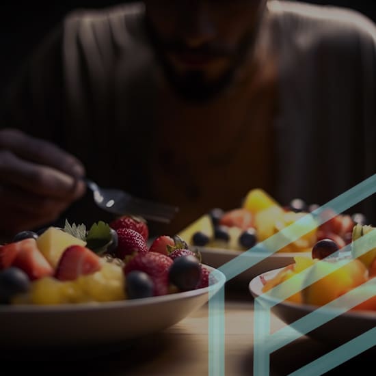 A person in dim lighting is preparing to eat from a bowl filled with various fruits, including strawberries, blueberries, and pineapple chunks. Another similar bowl sits nearby. The image has a semi-transparent geometric overlay on the right.