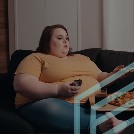 A person with brown hair wearing a yellow shirt and blue jeans sits on a dark sofa eating a slice of pizza. A drink and other food items can be seen on a table in front of them. The background includes a light-colored wall and a wooden accent.