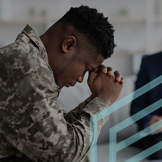 A person in military camouflage uniform sits with their head bowed and hands clasped in front of them, appearing contemplative or in prayer. They are indoors, with blurred objects in the background.