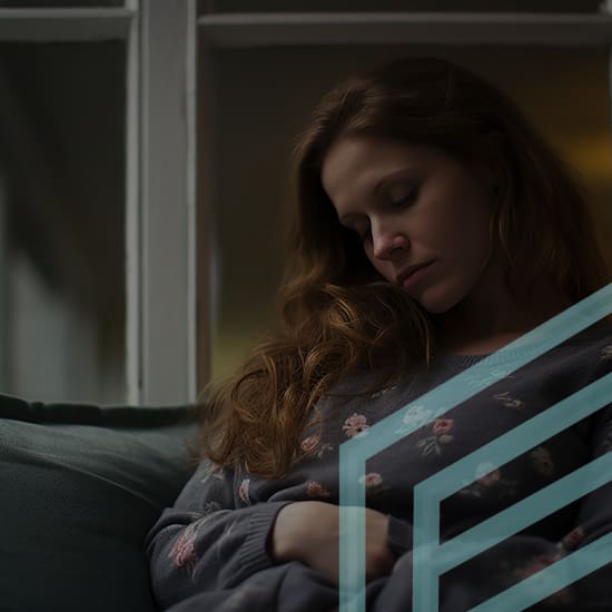 A woman with long hair, wearing a floral sweater, is seated on a sofa near a large window. She has her head tilted down and eyes closed, suggesting that she may be sleeping or deep in thought. The scene is dimly lit, creating a calm and introspective atmosphere.