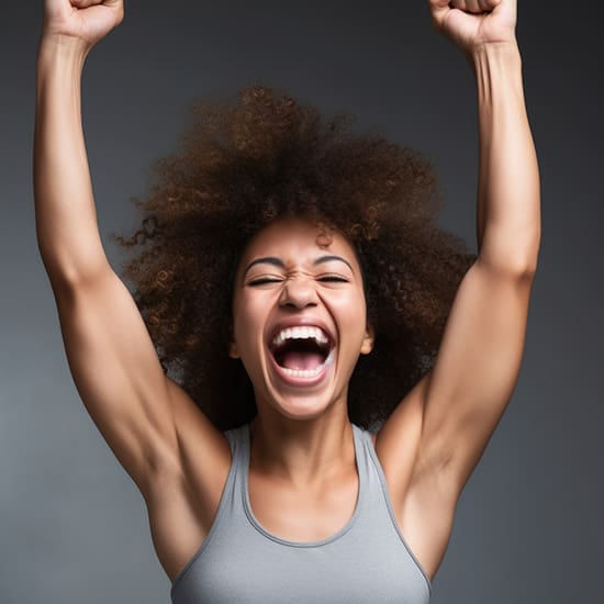 A person with curly hair is joyfully smiling and raising both arms overhead in a victorious pose. They are wearing a gray sleeveless top, and the background is a neutral gray.