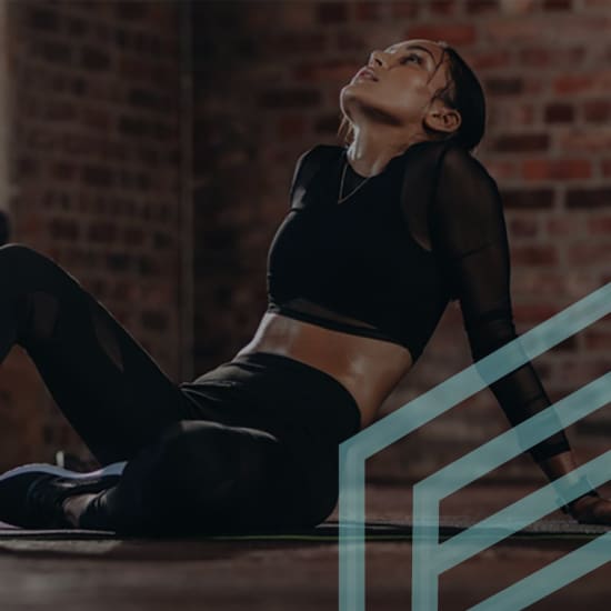 A woman in athletic wear with a sheer black long-sleeve top, sports bra, and leggings sits on the floor of a gym, leaning back on her hands, and looking upward. The background features exposed brick walls, and a geometric design overlays the image.