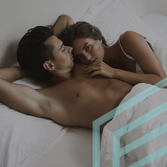 A couple lies in bed, with the man lying on his back and the woman snuggled up next to him, resting her head on his shoulder and touching his chest. They are covered partially by a white sheet. Soft light illuminates the scene, creating a serene and intimate atmosphere.