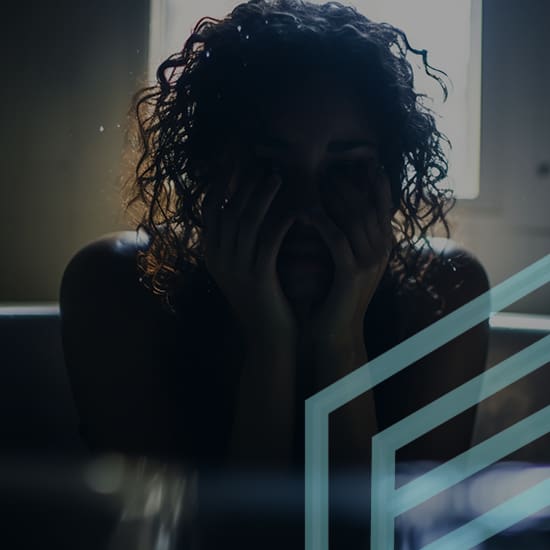 A person with curly hair is sitting in a dimly lit room, with hands covering their face in apparent distress. Light from a window behind them illuminates their silhouette, and diagonal transparent geometric shapes overlay the image.
