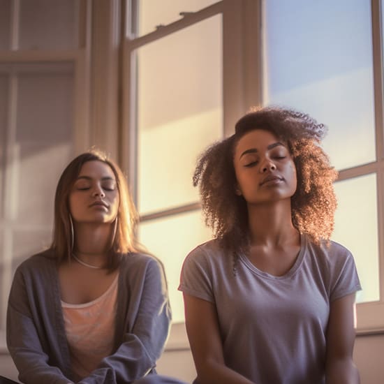 Two women sitting with eyes closed in a sunlit room, appearing to be meditating. The soft light from the large windows creates a calm and serene atmosphere. One woman has curly hair and the other has straight hair, both wearing casual clothing.