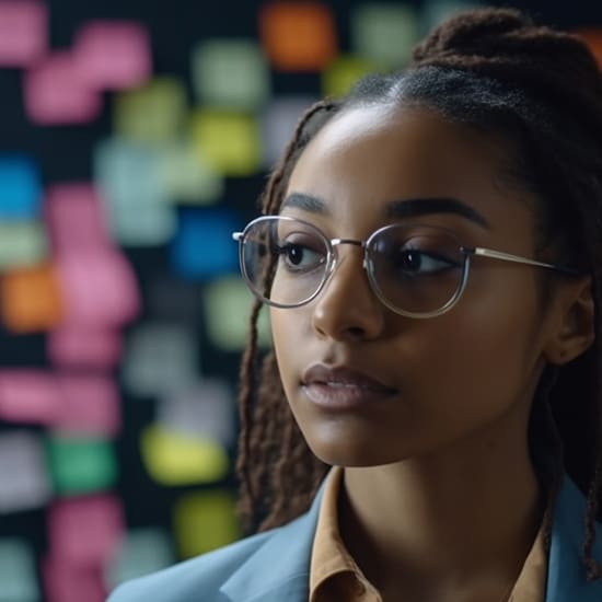 A person with glasses and dreadlocks is gazing thoughtfully to the side. In the background, there are colorful sticky notes on a board. They are wearing a light blue blazer and a tan collared shirt.