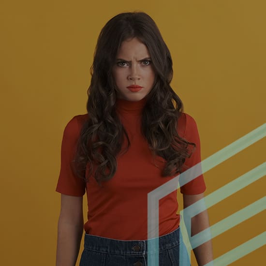 A woman with long, wavy brown hair and red lipstick stands against a yellow background, wearing a red turtleneck shirt and blue high-waisted jeans. She has an angry expression on her face. Light green geometric lines partially obscure her lower body.