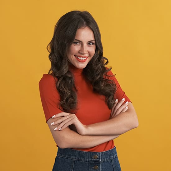 A woman with long, wavy, dark hair and a warm smile stands against a yellow background. She is wearing a red short-sleeved top and high-waisted denim jeans, with her arms crossed over her chest.