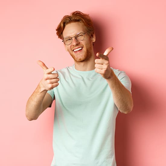 A smiling man with red hair and glasses, wearing a light green t-shirt, stands in front of a pink background. He is pointing both index fingers towards the camera in a friendly and playful manner.