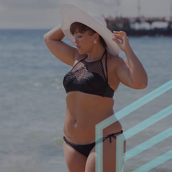 A woman wearing a black bikini and large white sunhat stands on a beach, with the ocean in the background. She holds the brim of her hat with one hand and has the other hand on her hip, gazing into the distance.
