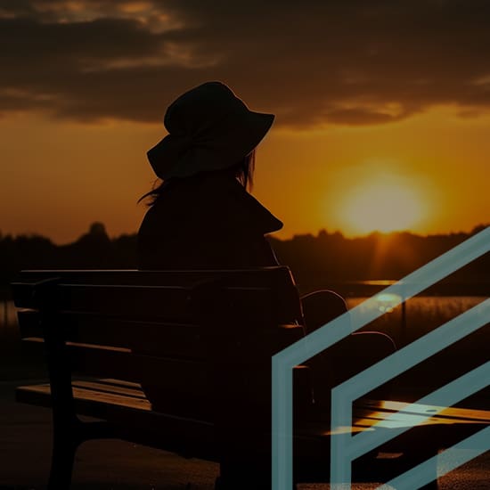 A person wearing a hat sits on a bench, facing a sunset over a body of water. The sky is painted with warm orange hues as the sun descends. Silhouettes of trees line the horizon, and geometric shapes overlay part of the scene in the foreground.