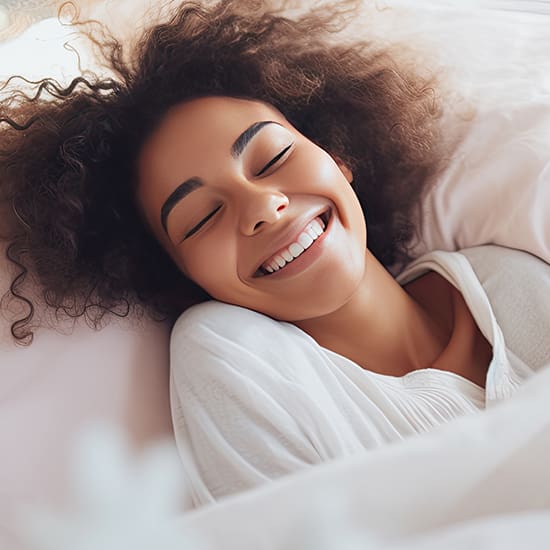 A person with curly hair smiles widely with eyes closed while lying in bed. They are wearing a white shirt and appear content and relaxed.