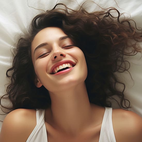 A woman with long, wavy hair lies on a white bed smiling widely with her eyes closed. She is wearing a white tank top and appears relaxed and happy.