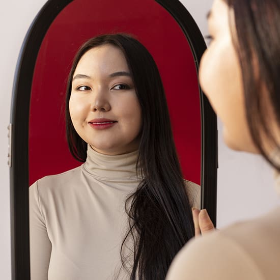 A woman with long dark hair and a beige turtleneck top is smiling and looking into a mirror, reflecting her face. The background of the mirror is red, contrasting with the white wall behind her.