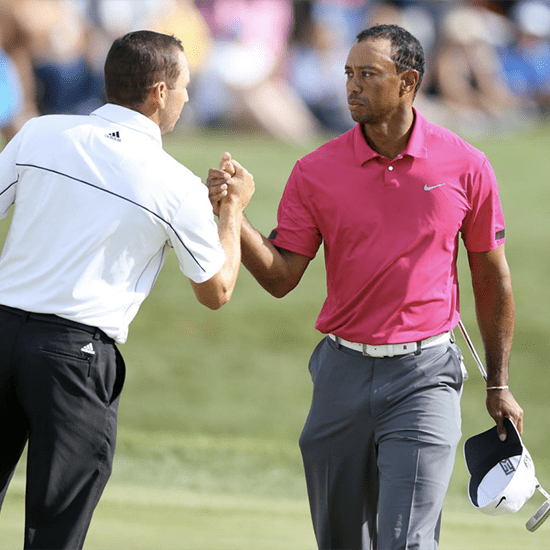 Two golfers are shaking hands on a golf course. One golfer is wearing a pink shirt and gray pants, holding a white cap in his left hand. The other golfer is dressed in a white shirt and black pants. A blurred crowd in the background watches the exchange.