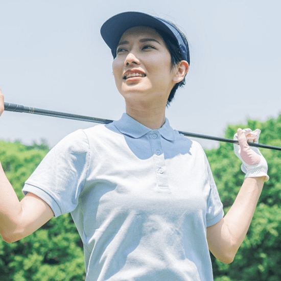 A woman stands outdoors on a golf course, holding a golf club over her shoulders. She is dressed in a light blue polo shirt and a navy visor. She is smiling and gazing off into the distance. Green foliage is visible in the background.