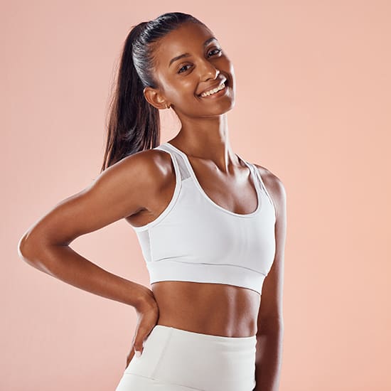 A person stands against a peach-colored background, smiling and looking at the camera. They have long, dark hair tied back into a ponytail and are wearing a white sports bra and matching leggings. The person's left hand rests on their hip.