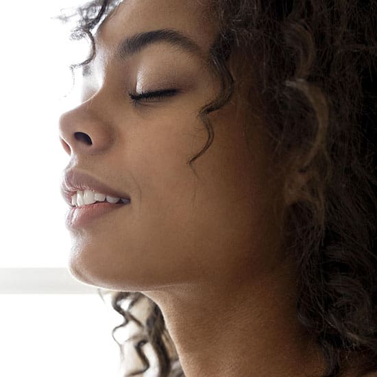 A close-up image of a person with curly hair and closed eyes, facing left. They have an expression of contentment or serenity. The background is softly lit, emphasizing the calm and peaceful mood of the scene.