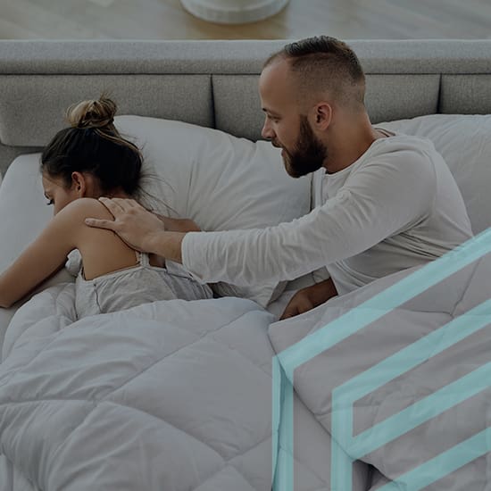 A man with a beard gently touches the back of a woman who is lying face down on a bed under a white blanket. Both are in a modern bedroom with light grey tones and minimal decor. The woman appears to be resting while the man offers comfort.