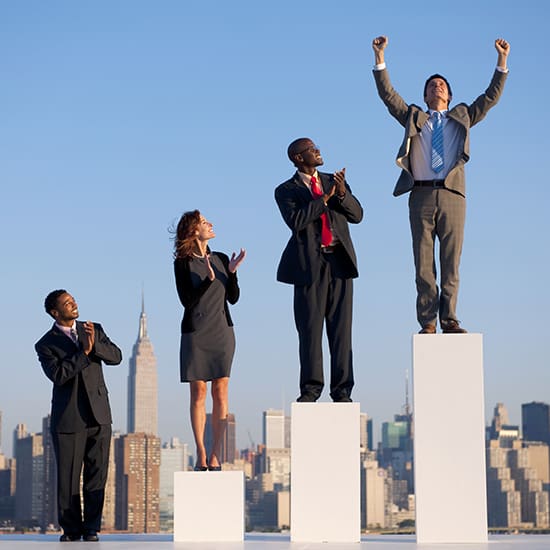 Four business professionals stand on different height podiums against a city skyline. The person on the tallest podium raises their arms in victory, the others applaud. Two men wear suits, one man and one woman wear business attire.