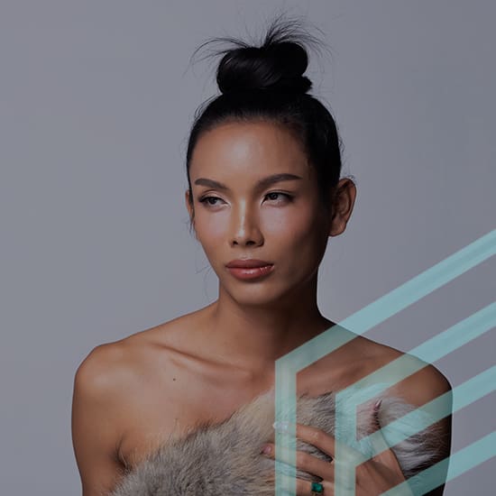 A person with a high bun poses against a gray background, draped in a fur-like material. They have a serene expression, with highlighted cheekbones and subtle makeup. Geometric light blue lines overlay the bottom right of the image.