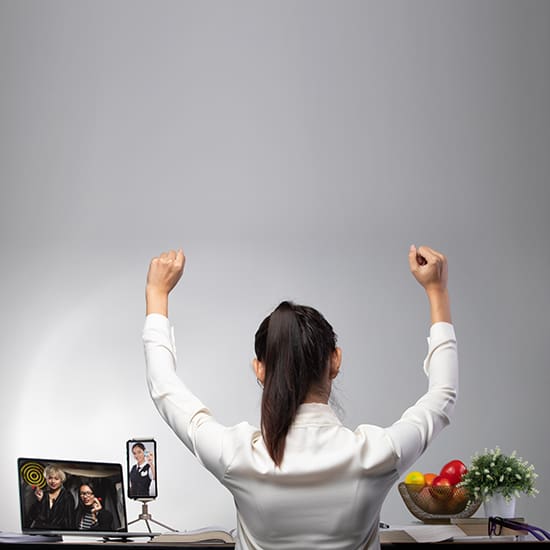 A person with a ponytail, wearing a white shirt, is seated at a desk with their back to the camera, raising both hands in the air. The desk has framed family photos, a basket of fruit, a small potted plant, and some electronic devices on it.