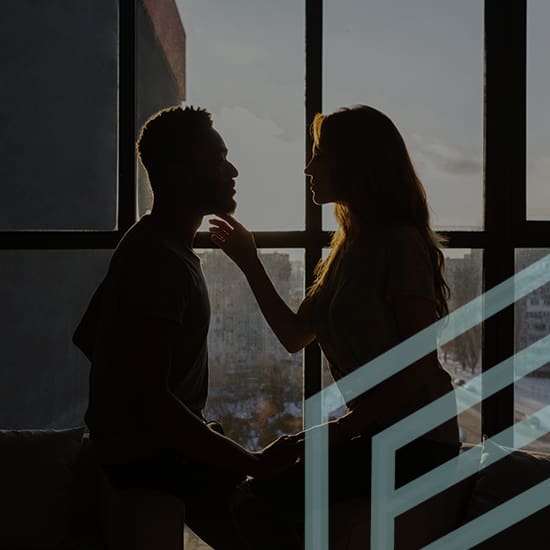 A silhouetted couple sits closely together by a large window, with the woman gently touching the man's chin. The scene is set during sunset or sunrise, casting a soft light on them. A geometric design is overlaid in the foreground.