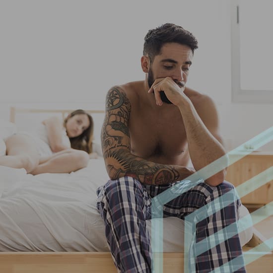 A shirtless man with tattoos and wearing plaid pants sits on the edge of a bed looking thoughtful. A woman lies on the bed in the background, partially covered by a blanket, gazing at him. The bedroom is simple with wooden furniture and natural light.
