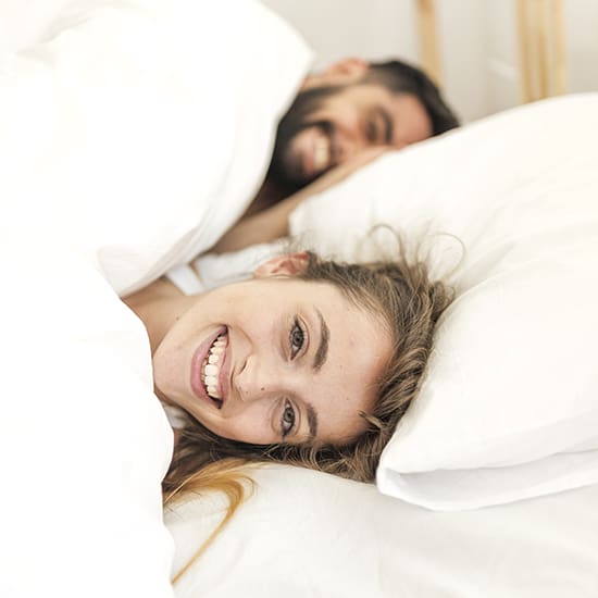 A woman smiles while lying in bed under a white duvet, with her head resting on a white pillow. In the background, a man, also lying in bed, smiles as well. Both appear relaxed and happy in the cozy setting.