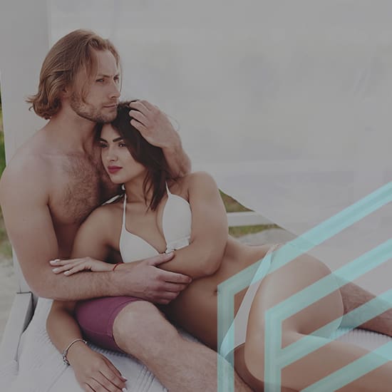 A man and woman in swimwear sit closely together on a white lounge bed. The man has his arm around the woman's shoulder, and they appear relaxed and content. The scene is bright, and there's a hint of outdoor scenery in the background.