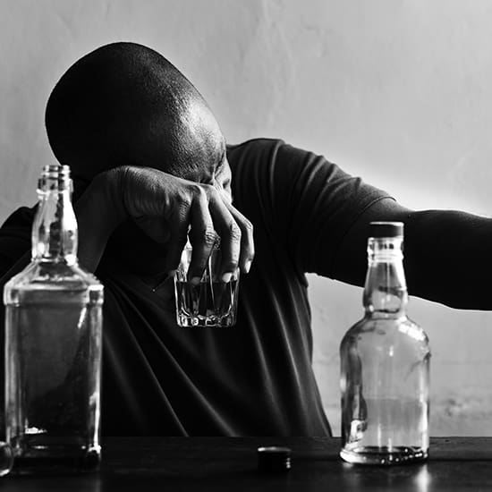 A person sits at a table with several empty and half-empty bottles of alcohol. They are holding a glass in one hand and have their head resting on their other arm, appearing distraught or exhausted. The image is black and white.