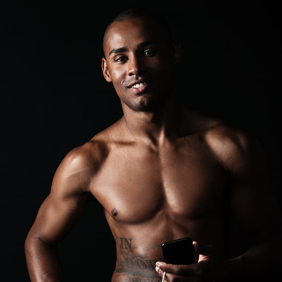 A muscular, shirtless man with short hair stands against a dark background, smiling slightly while holding a smartphone. The light accentuates his toned physique and tattoos are visible on his chest and lower abdomen.