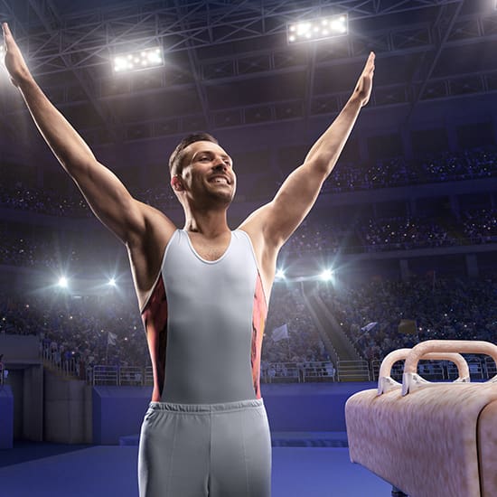 A male gymnast, wearing a white and red sleeveless leotard and white pants, stands with his arms raised in triumph. He appears to be in a large indoor arena filled with an audience, with bright spotlights shining from above. An apparatus is visible beside him.