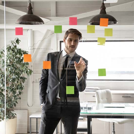 A man in a suit stands behind a glass wall covered with colorful sticky notes, pointing at one of the notes. He is in a modern office setting with large windows, a potted plant, and a desk visible in the background.