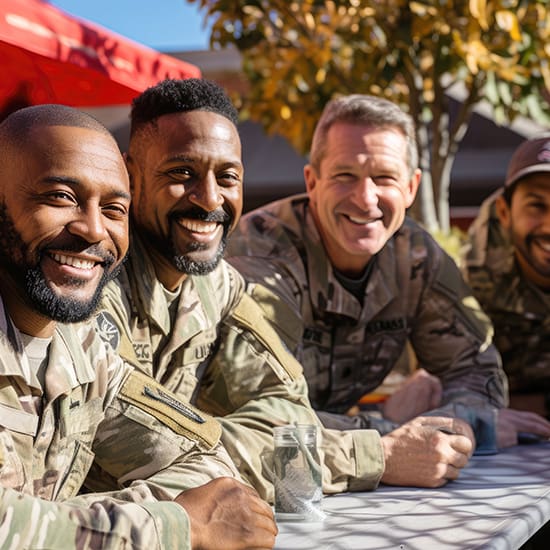 Four soldiers in uniform are smiling and leaning on a table outdoors on a sunny day. They appear relaxed and happy, with trees and a bright sky in the background. The group includes diversity in its members and conveys camaraderie and enjoyment.