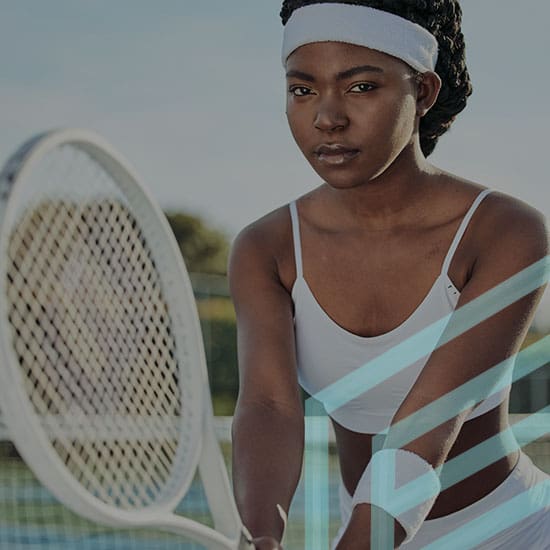 A tennis player in a white sports bra and headband holds a tennis racket, standing on a tennis court. The player appears focused and ready to hit the ball, with the blurred background showing parts of the court and greenery.