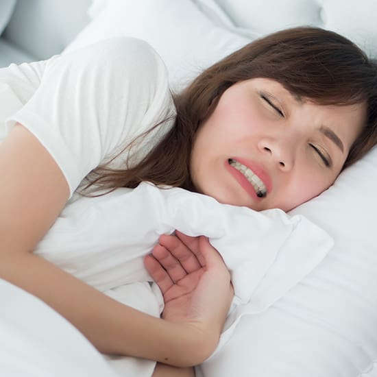A person with long brown hair lies in bed on a white pillow, appearing to be in discomfort or pain, with eyes closed and teeth clenched. One hand is tucked under the pillow while the other clutches part of the white blanket, a scene indicative of someone struggling with mental disorders. They wear a white shirt.