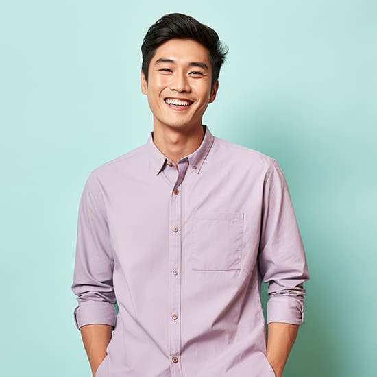 A smiling man stands in front of a light turquoise background, wearing a light purple button-up shirt with the sleeves rolled up. His hands are in his pockets, and he exudes a cheerful and relaxed vibe.